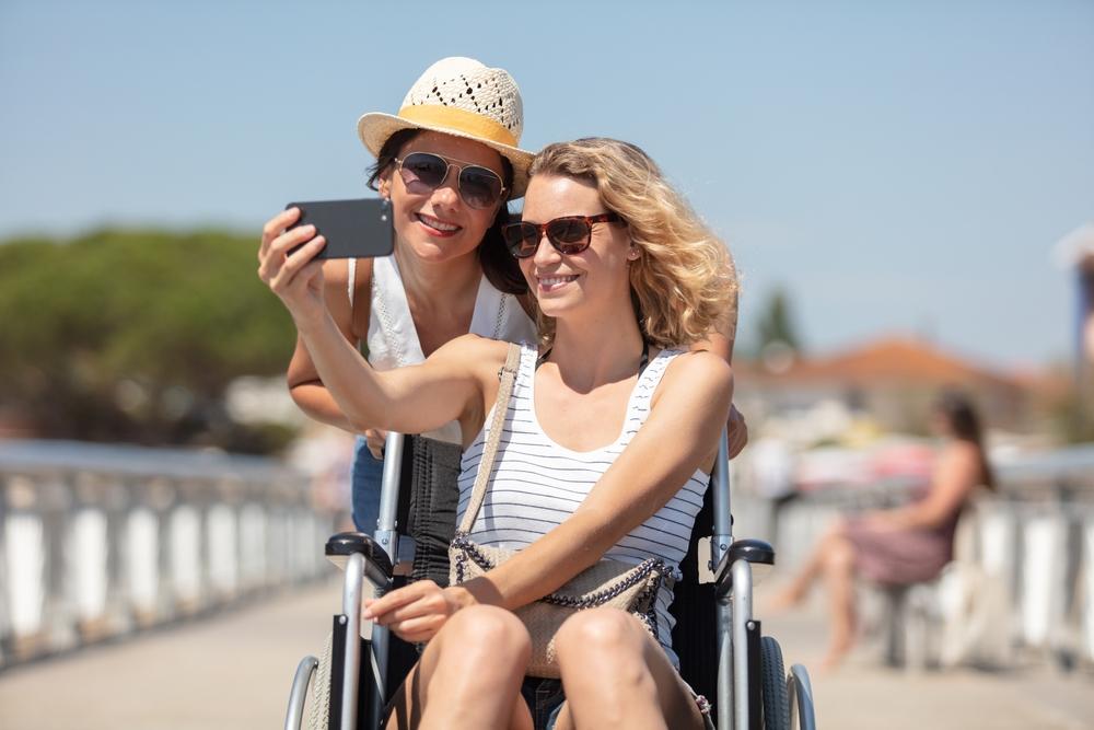 a woman in sunglasses sitting in a wheelchair taking a selfie with a friend